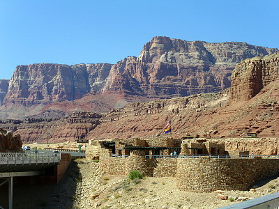 [Part of the pedestrian bridge railing is in the lower right of the image. Coming in from the left side is the vehicle bridge. In the center foreground is a stone structure with round wall sections between some straight sections. Encompassing the upper two thirds of the image are two sets of high cliff walls. The front one is mostly brown while the back one is different colors (red, brown, grey) in different sections of the depths of the rock. The cloudless sky is bright blue.]